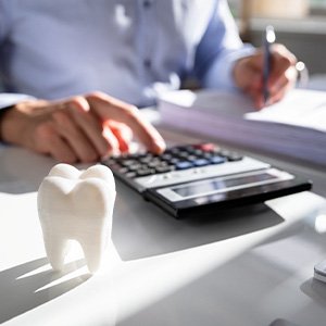 Someone calculating costs at desk with large model tooth, pen and paper, and calculator