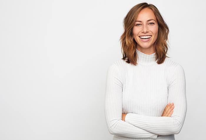 Woman in white sweater smiling in front of white background with arms crossed