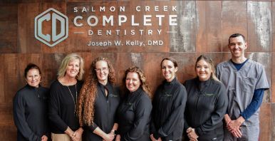 Dentist and team members smiling in front of wall with Salmon Creek Complete Dentistry logo